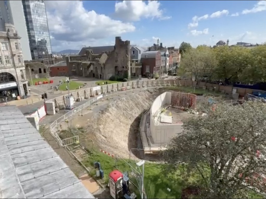 Wide view of the Castle Square Gardens excavation, with a curved concrete wall, site fencing and the ruins of Swansea Castle in the background