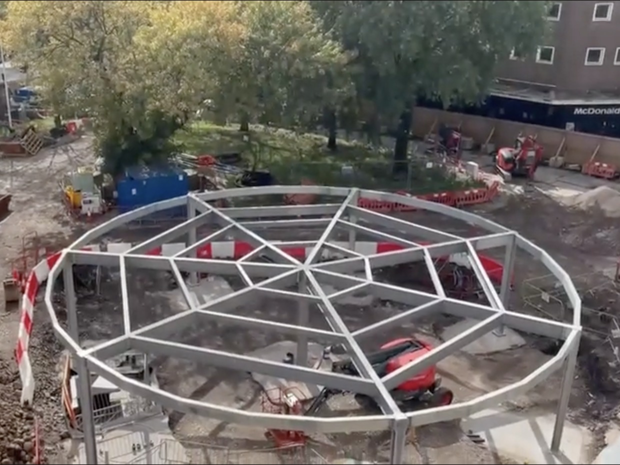 Circular steel frame of a pavilion under construction on the Castle Square Gardens site, with diggers and trees behind