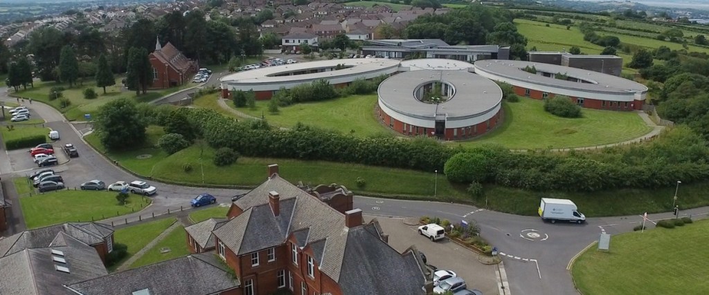 Aerial view of the modern circular red-brick and grey-roofed mental health unit at Cefn Coed Hospital, Swansea, with the older red-brick hospital building in the foreground and housing beyond