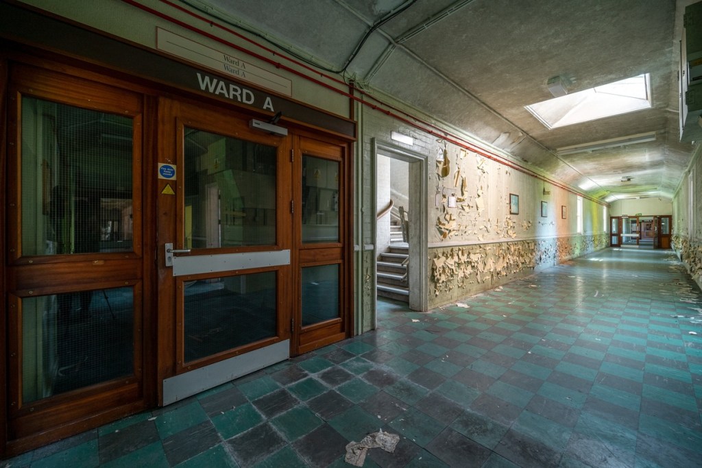 A long empty hospital corridor with peeling cream paint, a green and black chequered tiled floor and a set of wooden double doors marked Ward A