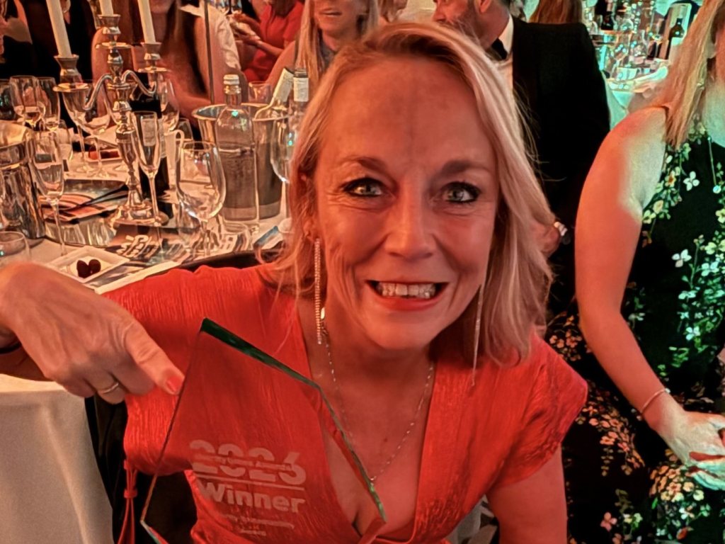 Cherrie Bija in a red dress, smiling at a dinner table and pointing to a glass trophy etched 2026 Winner