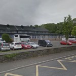 Two coaches and college minibuses parked in front of the long low Coleg Sir Gâr building in Ammanford, with cars in the car park and a stone wall by the road