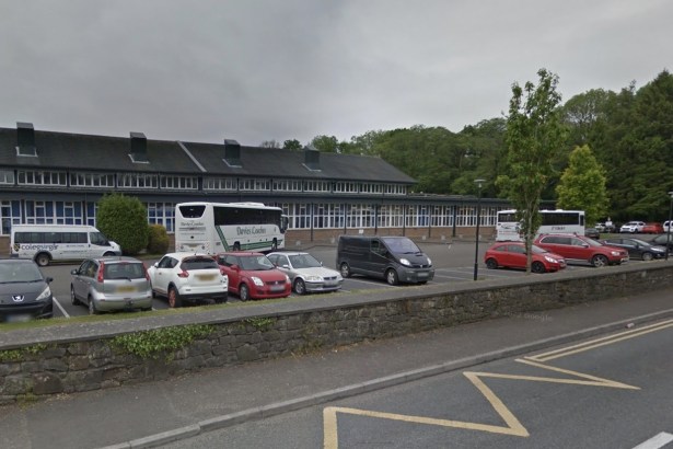 Two coaches and college minibuses parked in front of the long low Coleg Sir Gâr building in Ammanford, with cars in the car park and a stone wall by the road