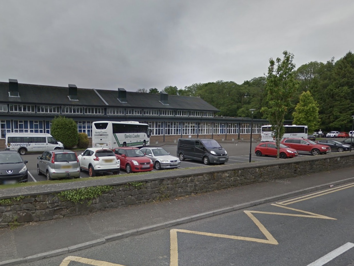 Two coaches and college minibuses parked in front of the long low Coleg Sir Gâr building in Ammanford, with cars in the car park and a stone wall by the road