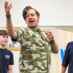 Stefan Pejic, in a camouflage shirt, gesturing mid-speech in a rehearsal room with two cast members behind him