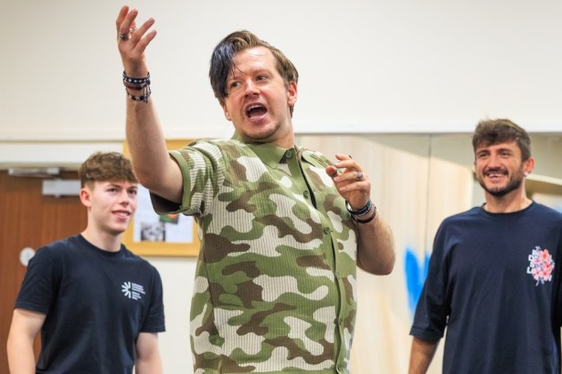 Stefan Pejic, in a camouflage shirt, gesturing mid-speech in a rehearsal room with two cast members behind him