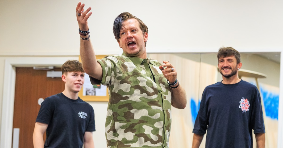 Stefan Pejic, in a camouflage shirt, gesturing mid-speech in a rehearsal room with two cast members behind him