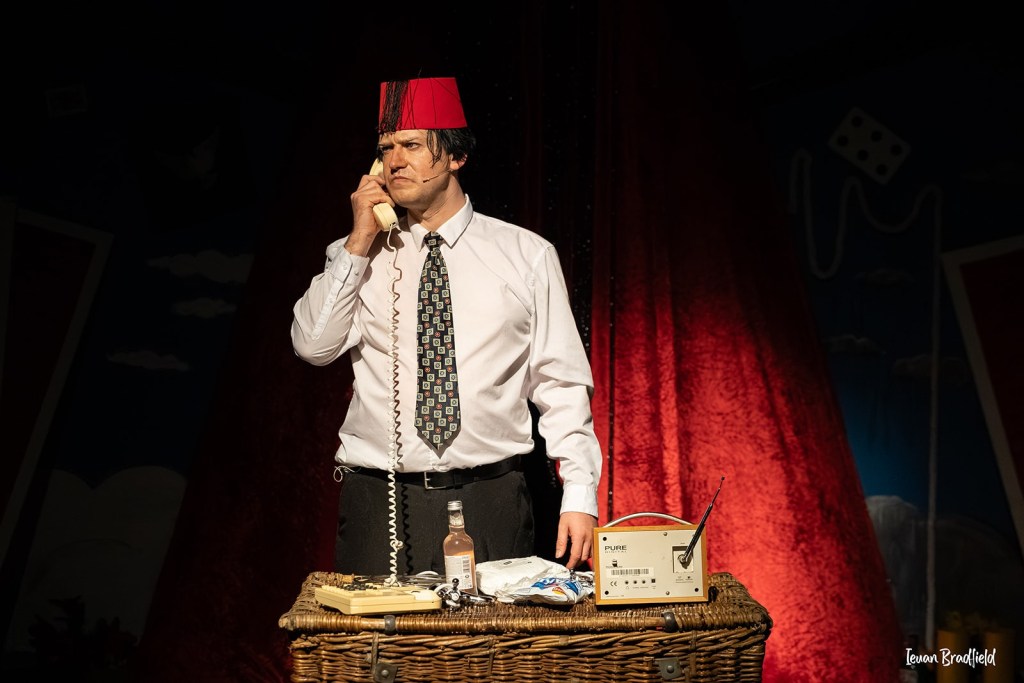 A performer in a red fez, white shirt and patterned tie holds a telephone receiver to his ear behind a wicker hamper with a bottle and a radio on it, against a red curtain