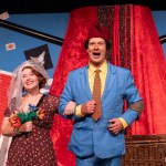 A performer in a red fez and bright blue suit links arms with a smiling woman in a bridal veil holding a bouquet, in front of a giant red fez and playing card set