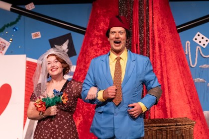 A performer in a red fez and bright blue suit links arms with a smiling woman in a bridal veil holding a bouquet, in front of a giant red fez and playing card set