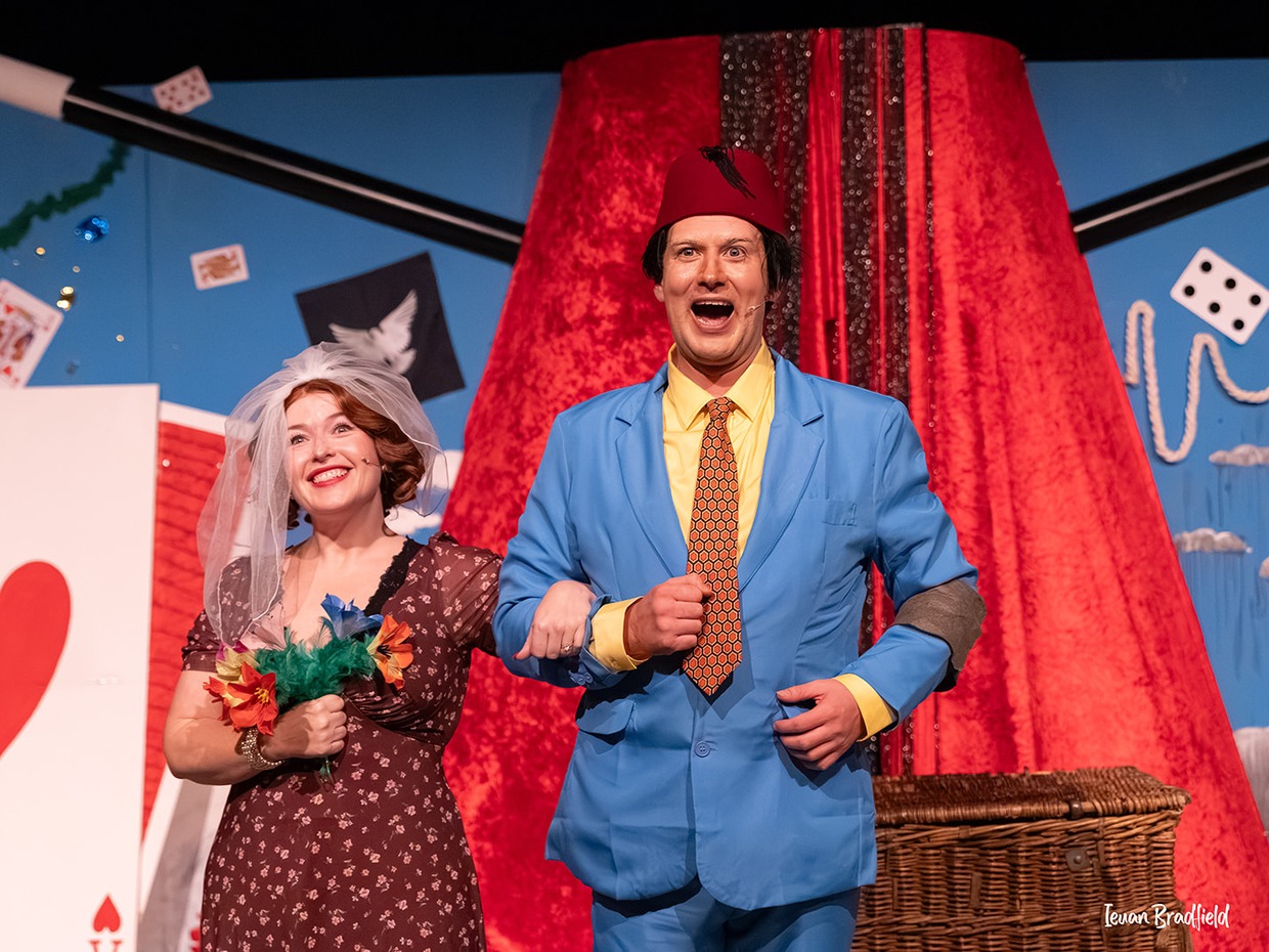 A performer in a red fez and bright blue suit links arms with a smiling woman in a bridal veil holding a bouquet, in front of a giant red fez and playing card set