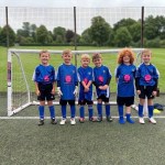 Six young children in blue football shirts, black shorts and blue socks stand smiling in a row in front of a small goal on an artificial pitch