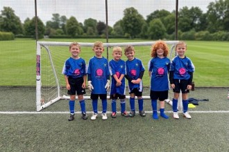 Six young children in blue football shirts, black shorts and blue socks stand smiling in a row in front of a small goal on an artificial pitch