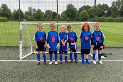 Six young children in blue football shirts, black shorts and blue socks stand smiling in a row in front of a small goal on an artificial pitch