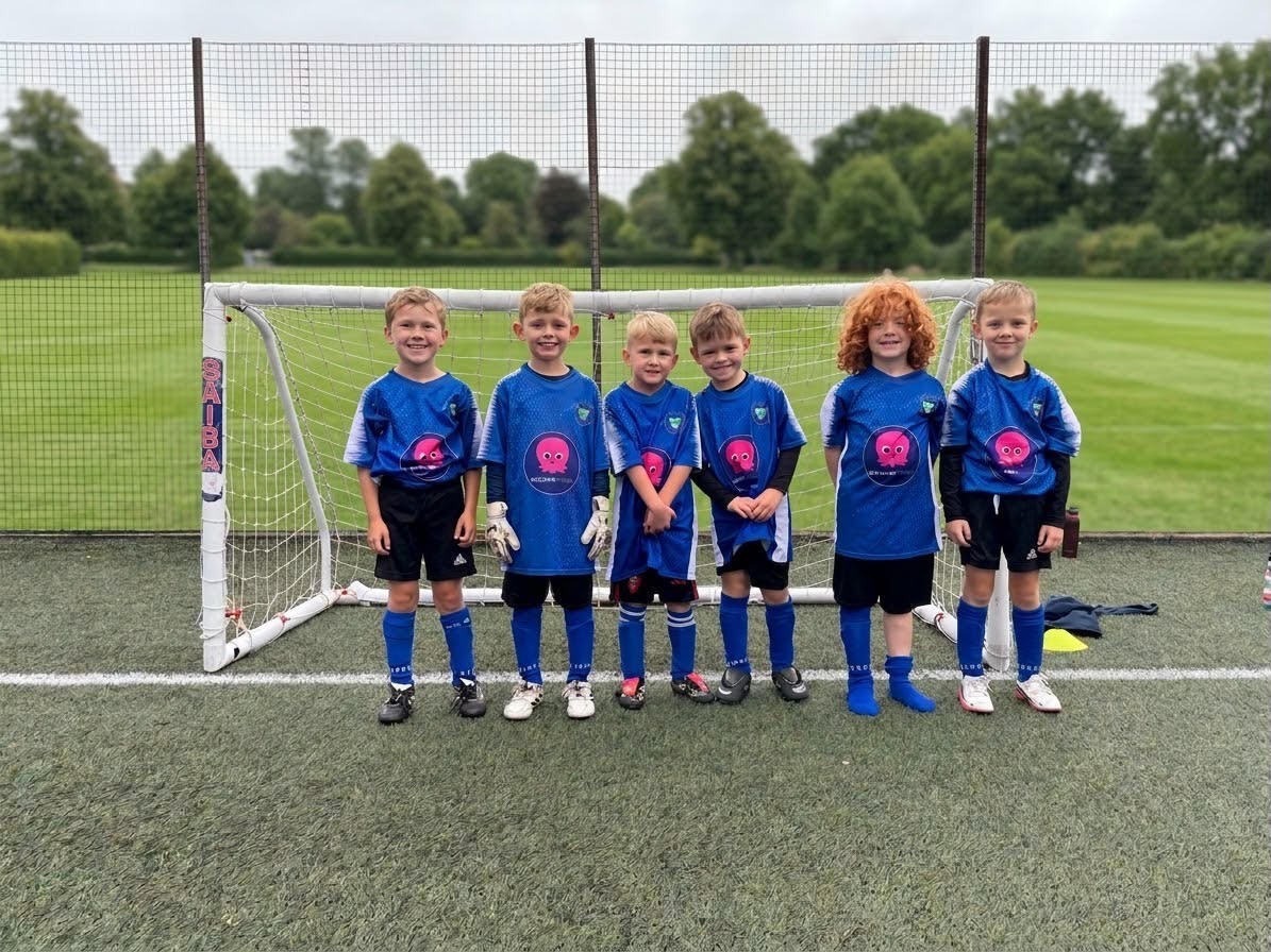 Six young children in blue football shirts, black shorts and blue socks stand smiling in a row in front of a small goal on an artificial pitch