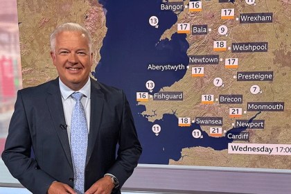 Derek Brockway in a dark suit and pale blue tie smiling beside a BBC Wales weather map showing temperatures across Wales