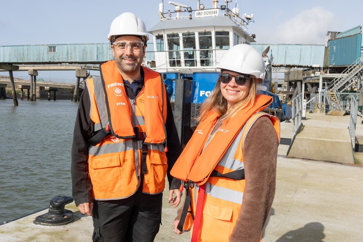 Diego Miguez and Marie Kelly in hard hats and orange life jackets on the quayside at Pembroke Dock with a vessel behind them