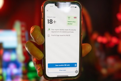 A hand holding a phone in a bar showing a digital proof of age screen reading 18+ and valid to buy alcohol