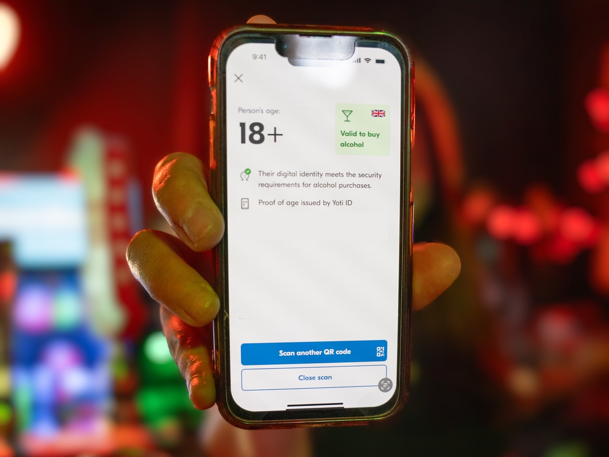 A hand holding a phone in a bar showing a digital proof of age screen reading 18+ and valid to buy alcohol