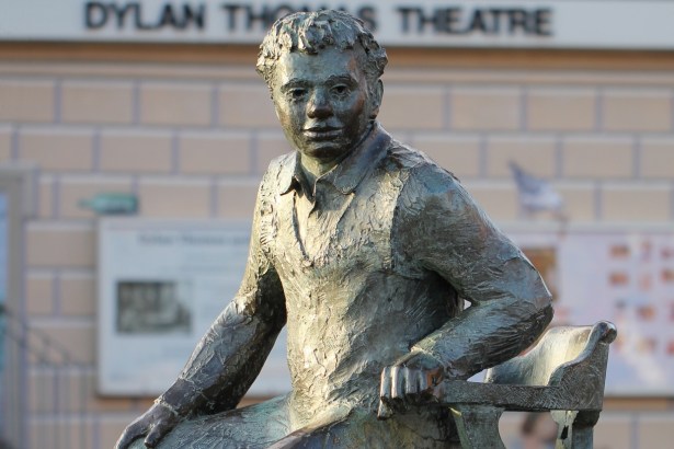 Bronze statue of Dylan Thomas seated on a chair, with the Dylan Thomas Theatre sign behind