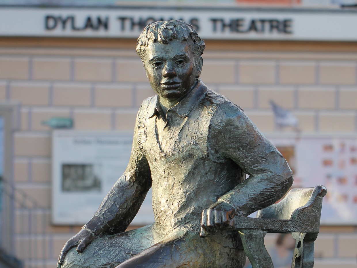Bronze statue of Dylan Thomas seated on a chair, with the Dylan Thomas Theatre sign behind