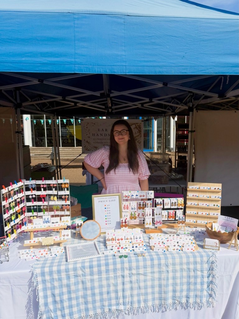 Ellie Jones under a blue gazebo at her earring stall, with the Swansea Museum Tramway Centre behind her