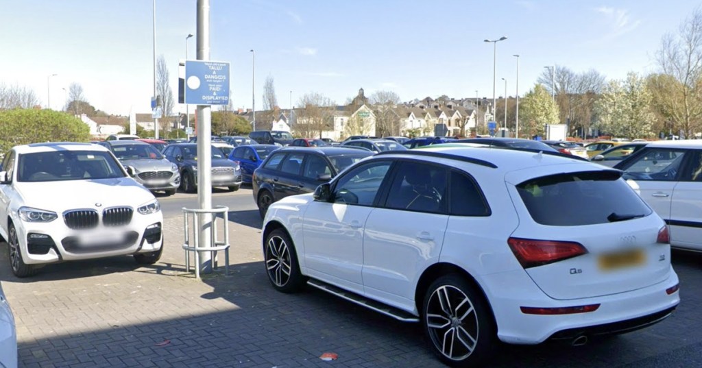 Parked cars at Eastgate car park in Llanelli beneath a sign reminding drivers to pay and display, with terraced houses beyond