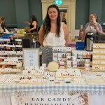 Ellie Jones standing behind her market stall of polymer clay earrings on a blue gingham cloth, with an Ear Candy Handmade UK banner and a price list