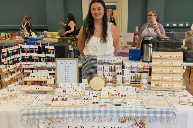 Ellie Jones standing behind her market stall of polymer clay earrings on a blue gingham cloth, with an Ear Candy Handmade UK banner and a price list