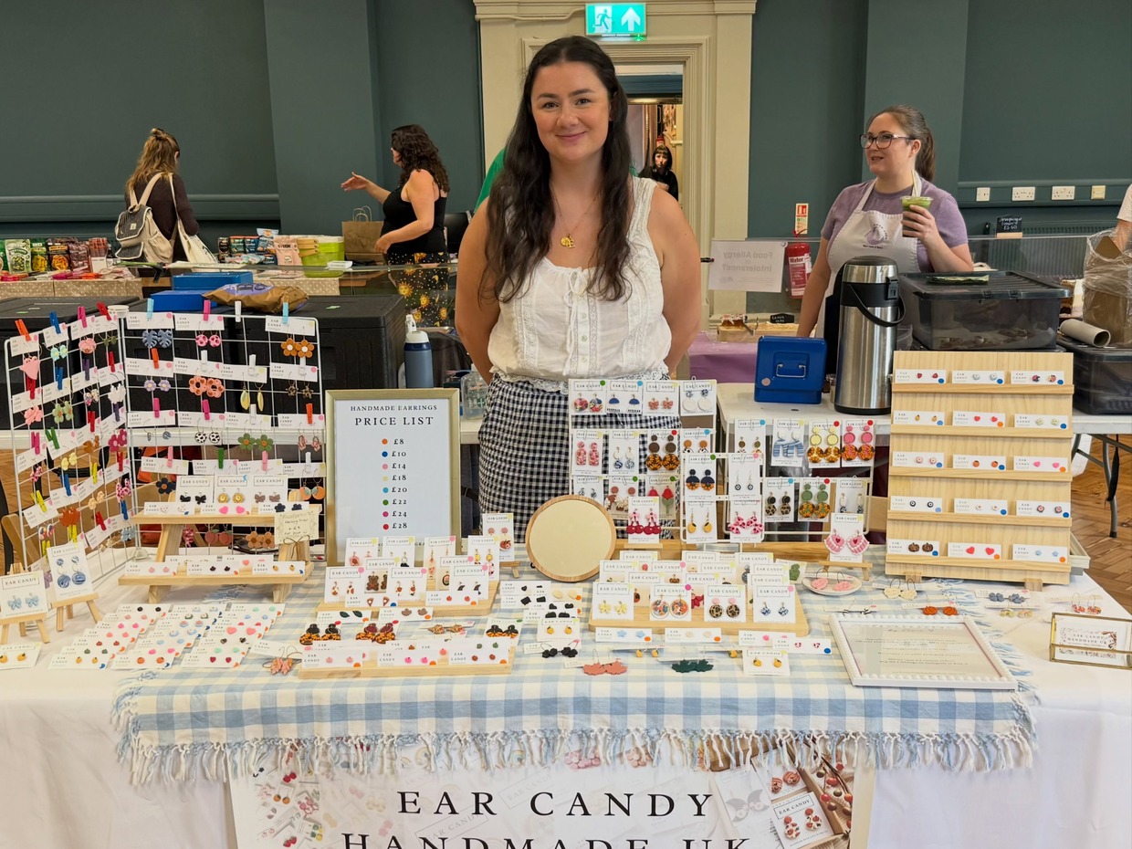 Ellie Jones standing behind her market stall of polymer clay earrings on a blue gingham cloth, with an Ear Candy Handmade UK banner and a price list