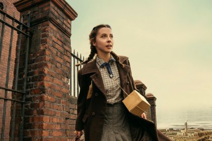 Erin Dolan as evacuee Rosie Birch, carrying a suitcase and gas mask box with an evacuation label on her coat, with Swansea's Guildhall tower and the bay behind her
