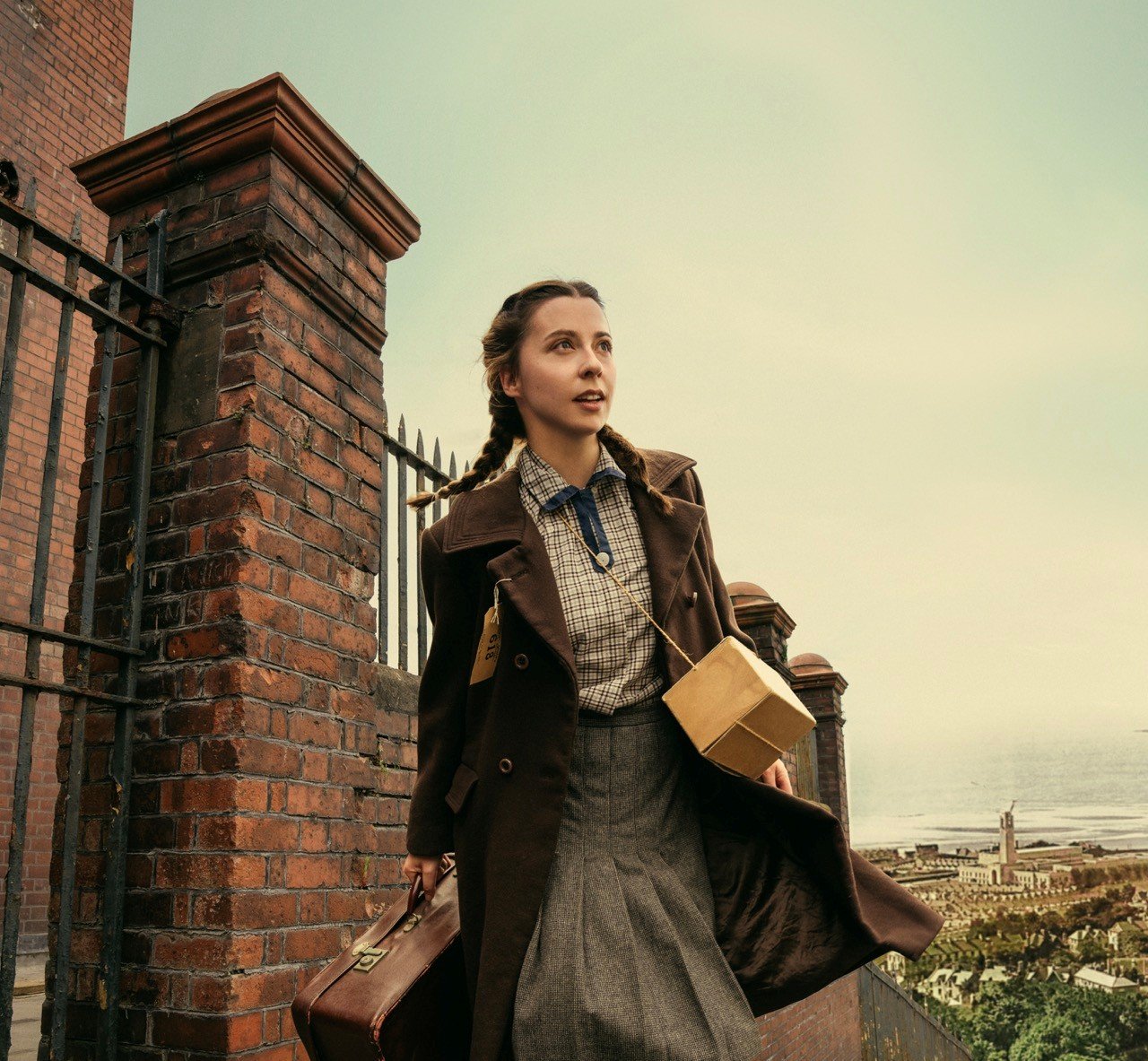Erin Dolan as evacuee Rosie Birch, carrying a suitcase and gas mask box with an evacuation label on her coat, with Swansea's Guildhall tower and the bay behind her