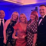 Five members of the Faith in Families team in evening wear under purple lighting, with chief executive Cherrie Bija in the centre holding a glass Charity Times Awards trophy