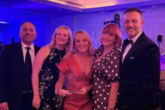 Five members of the Faith in Families team in evening wear under purple lighting, with chief executive Cherrie Bija in the centre holding a glass Charity Times Awards trophy