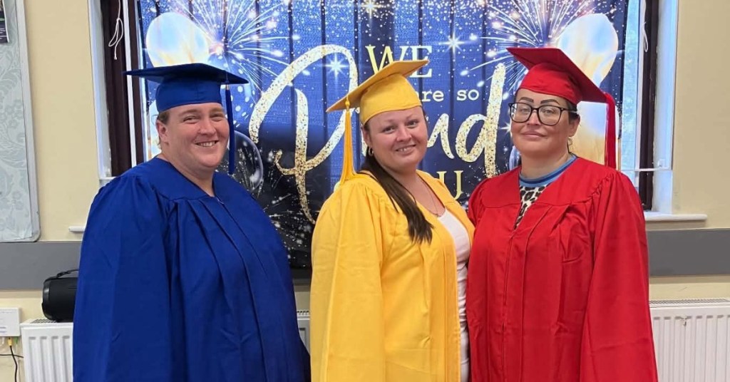 Three women in blue, yellow and red graduation caps and gowns smiling in front of a celebration banner at a Community Cwtch