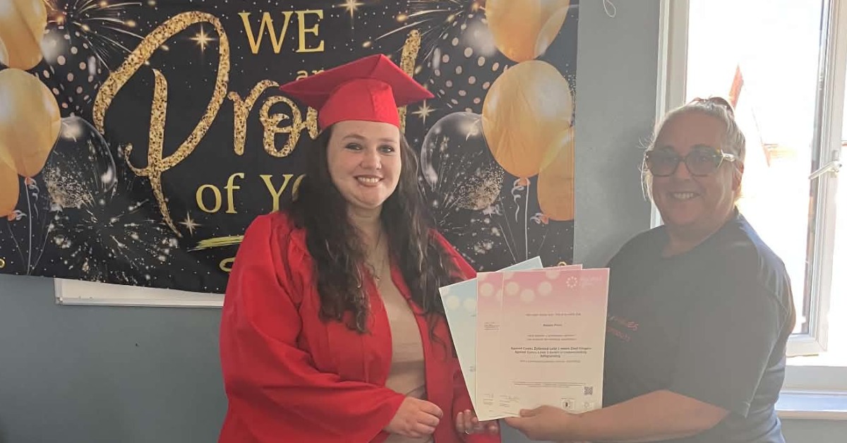 Natalie Price Pettersson in a red graduation cap and gown holding her certificates beside a Faith in Families staff member, in front of a celebration banner