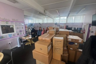 Dozens of cardboard boxes of school uniform stacked in a school community room, with staff standing behind them