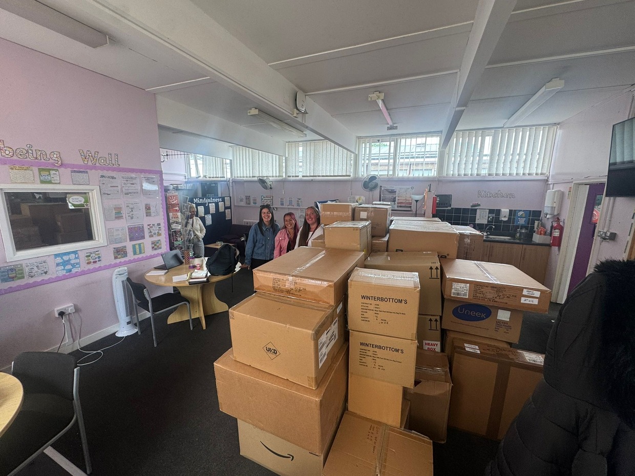 Dozens of cardboard boxes of school uniform stacked in a school community room, with staff standing behind them