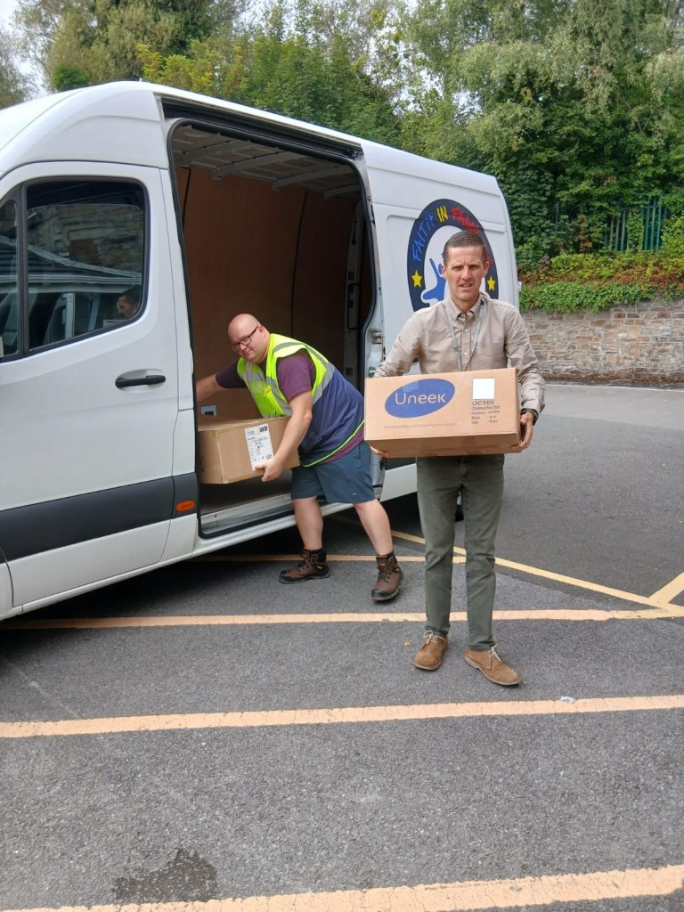 Two men unloading boxes of school uniform from a Faith in Families charity van in a school car park