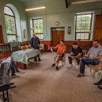 The Fflamau Penyberth company in rehearsal in a chapel vestry, seated in a circle with a keyboard and sheet music