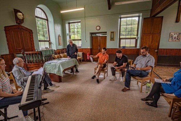 The Fflamau Penyberth company in rehearsal in a chapel vestry, seated in a circle with a keyboard and sheet music