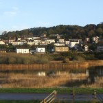 A reed-fringed lake reflecting the houses of Pwll, which climb a wooded hillside behind it, with the Wales Coast Path in the foreground