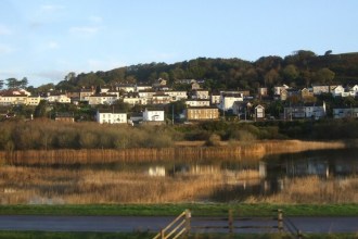 A reed-fringed lake reflecting the houses of Pwll, which climb a wooded hillside behind it, with the Wales Coast Path in the foreground