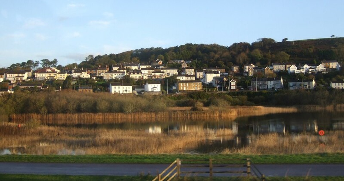 A reed-fringed lake reflecting the houses of Pwll, which climb a wooded hillside behind it, with the Wales Coast Path in the foreground