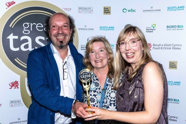 Three people smiling and holding a small gold fork-shaped Great Taste trophy in front of a sponsor board