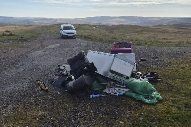 A pile of dumped household items including bags, bedding and a clothes airer on a gravel track across open hillside, with hills in the distance