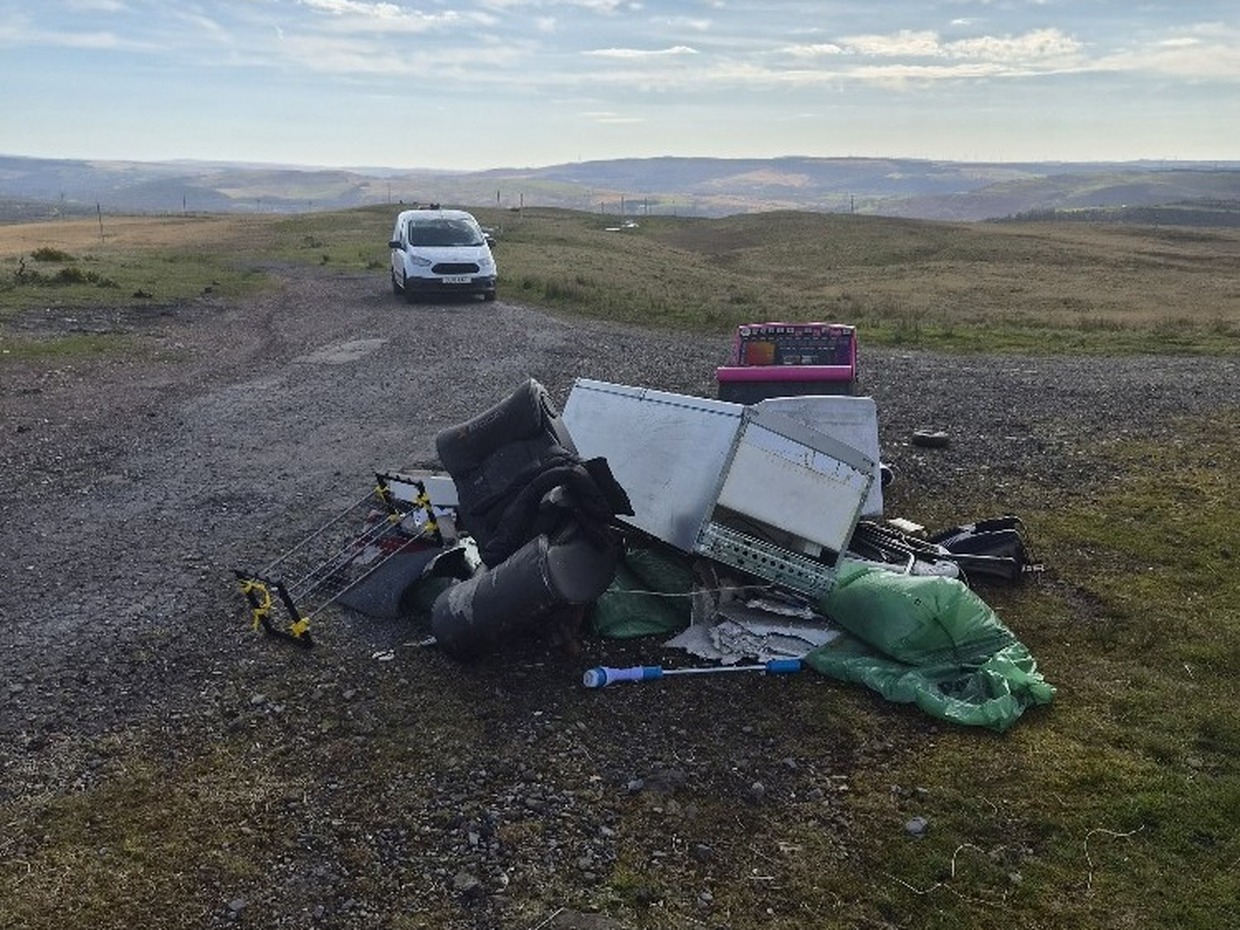 A pile of dumped household items including bags, bedding and a clothes airer on a gravel track across open hillside, with hills in the distance