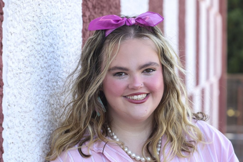 A close-up of Georgia Miggins smiling against a striped wall, wearing a pink hair bow, pearl necklace and pink striped shirt