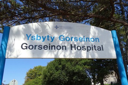 The bilingual Ysbyty Gorseinon Gorseinon Hospital entrance sign on blue posts beneath pine trees