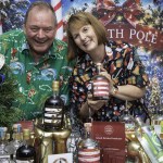 A smiling man and woman in Christmas shirts lean over a table of Gower Gin bottles in front of a North Pole backdrop, with a Small Drinks Producer of the Year award plaque